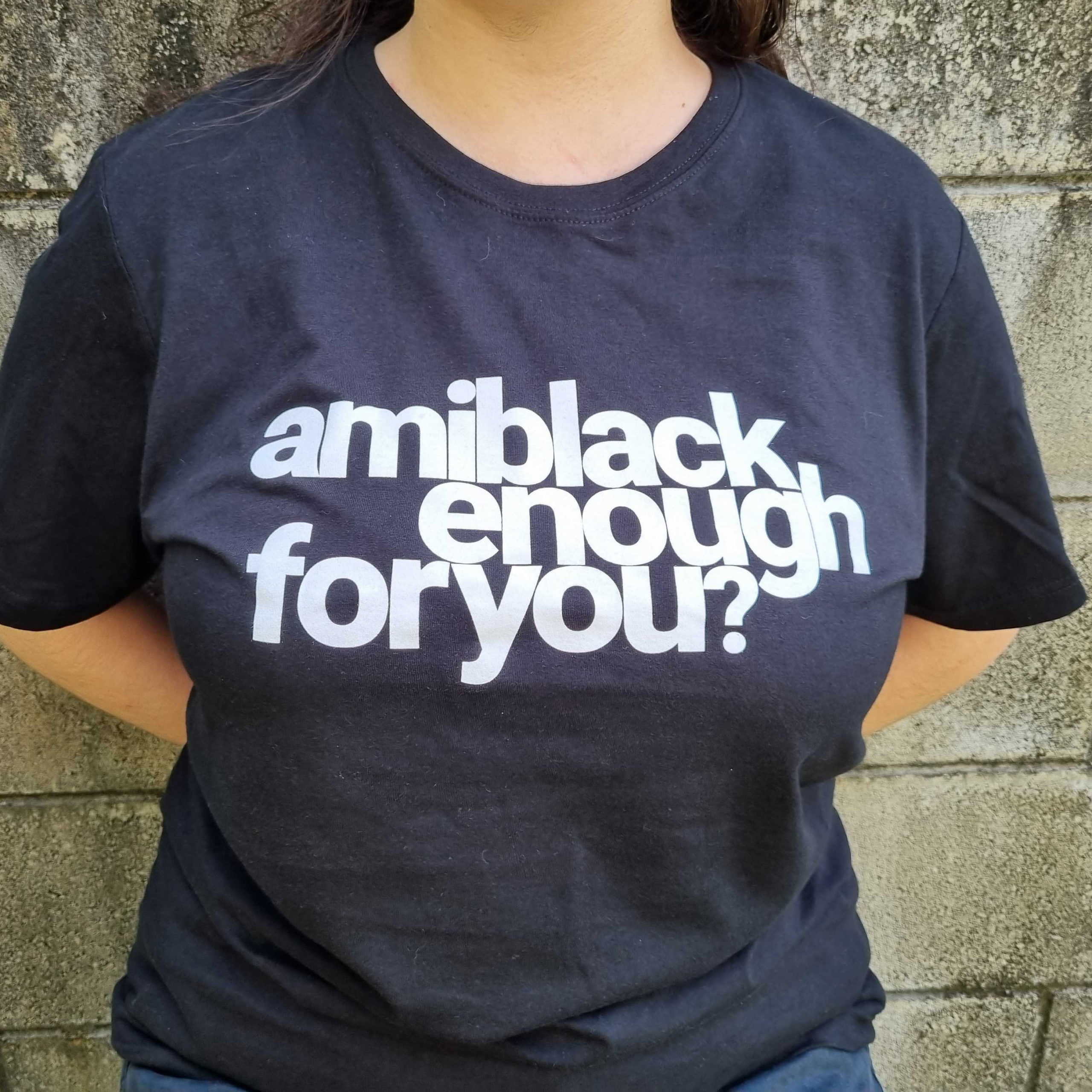 Against a concrete wall, a female person wearing a black tshirt with white bold text amiblackenoughforyou?